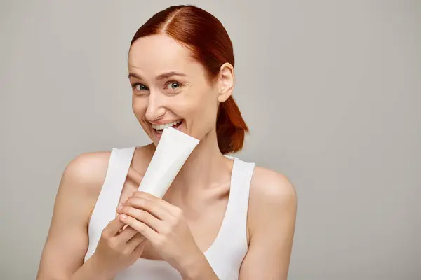 Cheerful woman with red hair biting tube with body lotion on grey background, skincare concept — Stock Photo