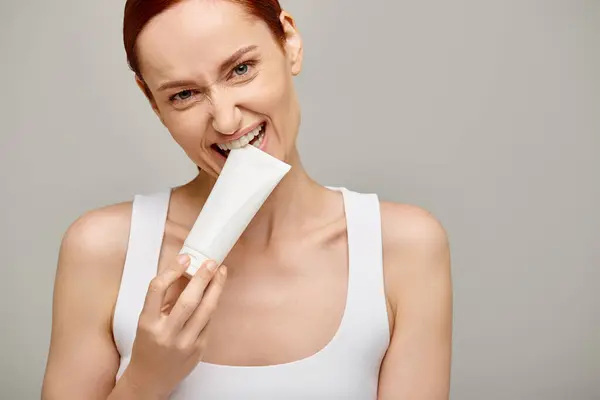Woman with red hair biting tube with lotion on grey background, skin and body care concept — Stock Photo
