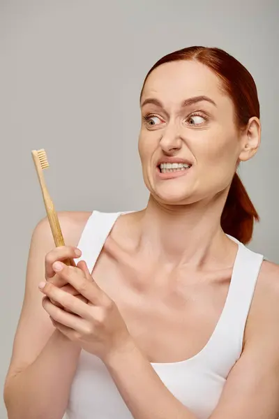 Femme rousse en débardeur grimaces à la brosse à dents en bois sur fond gris, concept d'hygiène dentaire — Photo de stock