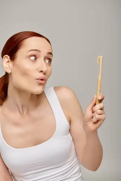 Mujer pelirroja sorprendida en camiseta sin mangas mirando el cepillo de dientes sobre fondo gris, concepto de higiene dental - foto de stock