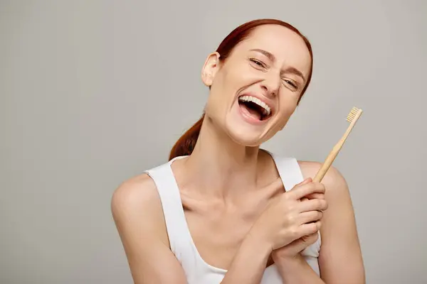 Happy redhead woman in tank top holding wooden toothbrush and smiling at camera on grey background — Stock Photo