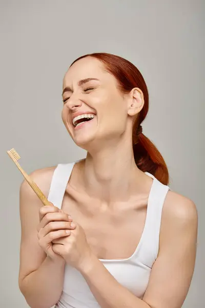 Happy redhead woman in tank top holding wooden toothbrush and smiling at camera on grey backdrop — Stock Photo