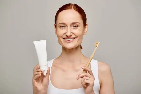Happy redhead woman in tank top holding toothpaste and toothbrush and smiling at camera on grey — Stock Photo