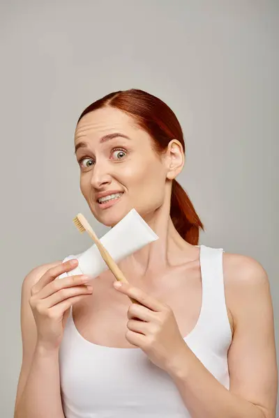 Confused redhead woman in tank top holding toothpaste and toothbrush and looking at camera on grey — Stock Photo