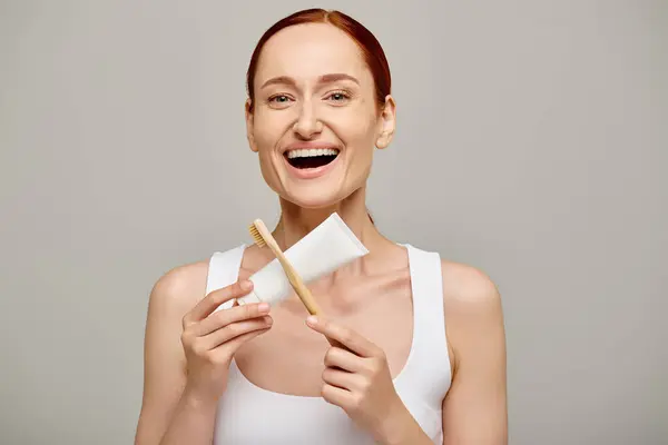 Femme rousse étonnée en débardeur tenant dentifrice et brosse à dents et souriant à la caméra sur gris — Photo de stock
