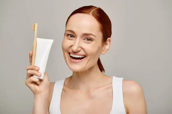 Aufgeregte Frau mit roten Haaren hält Zahnpasta und Zahnbürste in der Hand und lächelt in die Kamera auf grau — Stockfoto