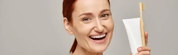 Aufgeregte Frau mit roten Haaren, die Zahnpasta und Zahnbürste in der Hand hält und auf grauem Banner in die Kamera lächelt — Stockfoto