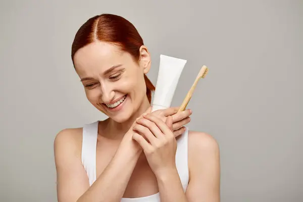 Fröhliche Frau mit roten Haaren, die Zahnpasta und Zahnbürste in der Hand hält und in die Kamera lächelt — Stockfoto