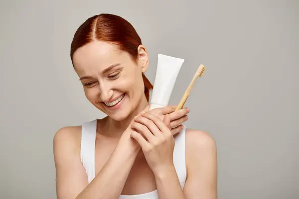 Joyful woman with red hair holding toothpaste and toothbrush and smiling at camera on grey — Stock Photo