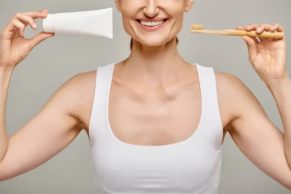 Cropped view of joyful woman holding toothpaste and toothbrush and smiling at camera on grey — Stock Photo