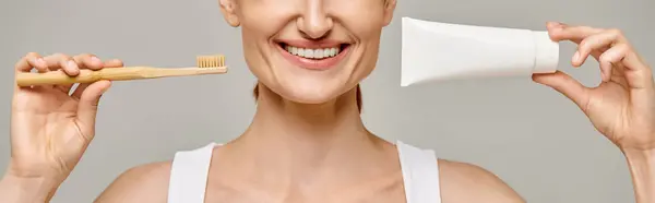 Cropped banner of joyful woman holding toothpaste and toothbrush and smiling at camera on grey — Stock Photo