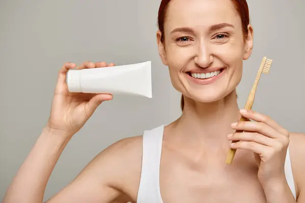 Happy woman with red hair holding toothpaste and toothbrush and smiling at camera on grey backdrop — Stock Photo