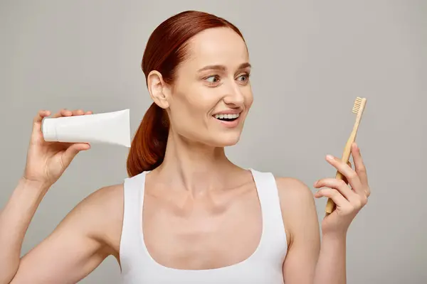 Staunte Frau mit roten Haaren, die Zahnpasta und Zahnbürste in der Hand hielt und vor grauem Hintergrund in die Kamera lächelte — Stockfoto
