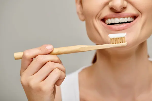 Ausgeschnittene Ansicht einer fröhlichen Frau, die Zahnbürste mit Zahnpasta in der Hand hält und auf grau in die Kamera lächelt — Stockfoto