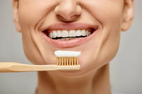 Cropped view of happy woman holding toothbrush with toothpaste and smiling at camera on grey — Stock Photo