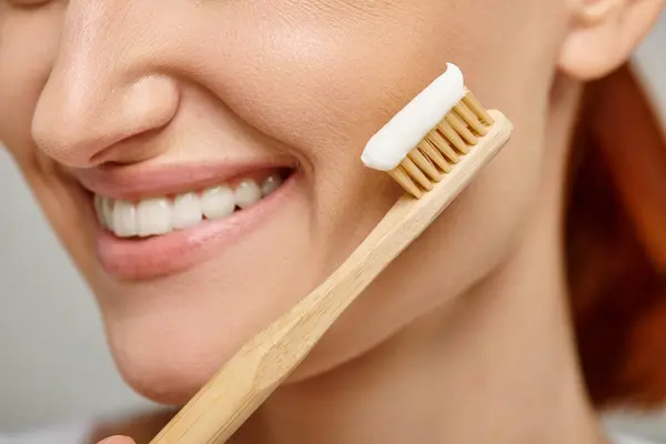 Cropped view of positive woman holding toothbrush with toothpaste and smiling at camera on grey — Stock Photo