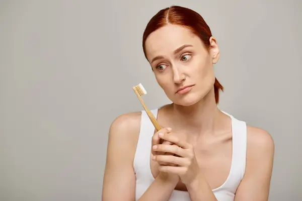 Vista recortada de mujer pelirroja sosteniendo cepillo de dientes con pasta de dientes sobre fondo gris, emoción - foto de stock