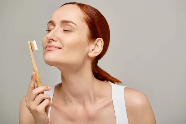 Mujer con pelo rojo sosteniendo cepillo de dientes de bambú con pasta de dientes sobre fondo gris, cuidado dental - foto de stock