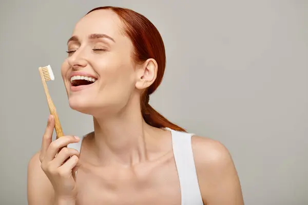 Excited woman with red hair holding bamboo toothbrush with toothpaste on grey backdrop, dental care — Stock Photo