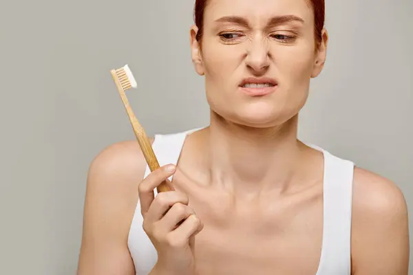 Displeased woman with red hair looking at bamboo toothbrush with toothpaste on grey backdrop, dental — Stock Photo