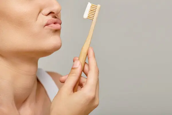 Displeased woman looking at bamboo toothbrush with toothpaste on grey backdrop, cropped photo — Stock Photo