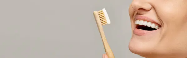 Vista recortada de mujer alegre sosteniendo cepillo de dientes con pasta de dientes y sonriendo sobre fondo gris. banner - foto de stock