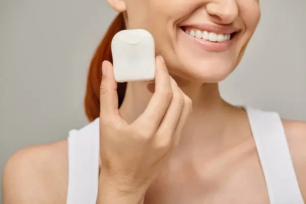 Cropped view of happy redhead woman holding dental floss case and smiling on grey background — Stock Photo