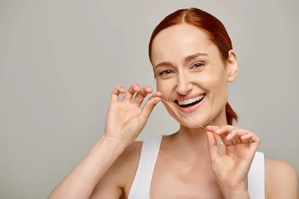 Heureuse rousse femme tenant fil dentaire et souriant sur fond gris, la promotion de l'hygiène buccodentaire — Photo de stock