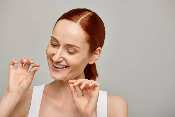 Fröhliche rothaarige Frau mit Zahnseide und Lächeln auf grauem Hintergrund, die Mundhygiene fördert — Stockfoto