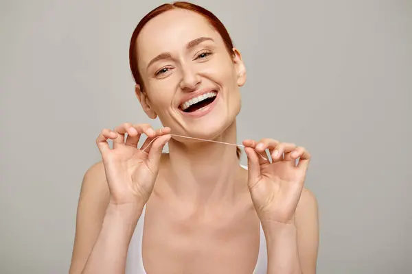 Excited redhead woman holding dental floss and smiling on grey background,  promoting oral hygiene — Stock Photo