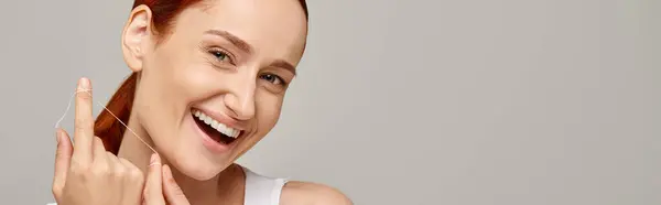 Aufgeregtes rothaariges Model mit Zahnseide und Lächeln auf grauem Hintergrund, Banner zur Mundhygiene — Stockfoto