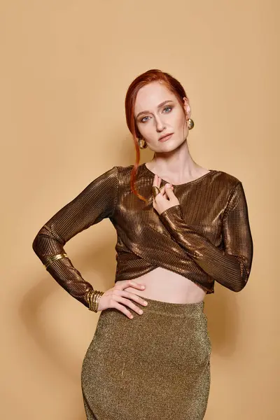 Redhead woman in her 30s posing in golden attire and accessories on beige backdrop, hand on hip — Stock Photo