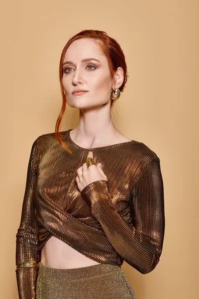 Elegant and redhead woman in her 30s posing in golden attire and accessories on beige backdrop — Stock Photo