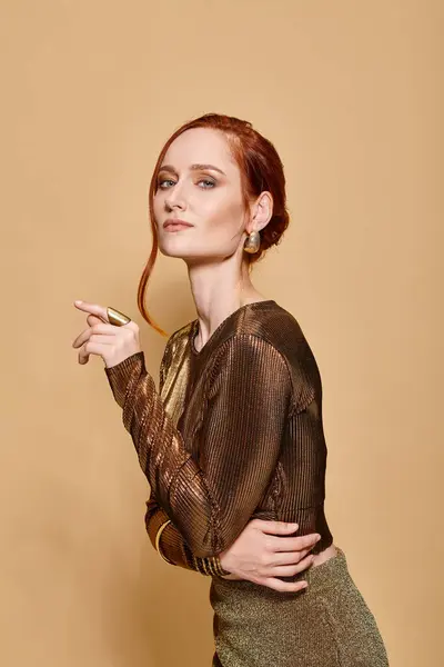 Graceful redhead woman in her 30s posing in elegant attire with golden accessories on beige backdrop — Stock Photo