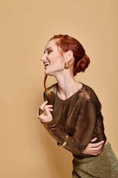 Rossa donna sulla trentina in posa in abito d'oro e accessori e sorridente sullo sfondo beige — Foto stock