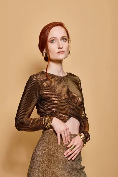 Anmutige rothaarige Frau in ihren Dreißigern posiert in trendiger Kleidung mit goldenen Accessoires auf beigem Hintergrund — Stockfoto