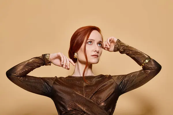 Dreamy woman in her 30s posing in stylish attire and golden accessories with raised hands on beige — Stock Photo