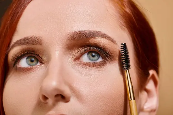 Cropped photo of redhead woman with green eyes holding eye brow brush on beige backdrop — Stock Photo