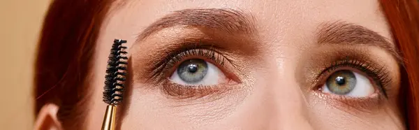 Cerrar foto de mujer pelirroja con ojos verdes aplicando rímel sobre fondo beige, pancarta - foto de stock