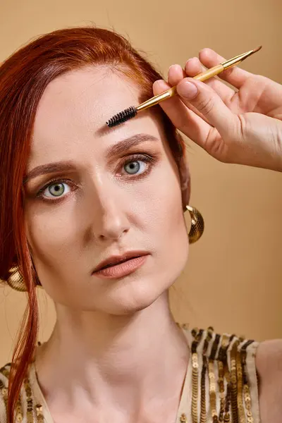 Portrait de femme rousse aux yeux verts appliquant mascara sur fond beige, maquillage — Photo de stock