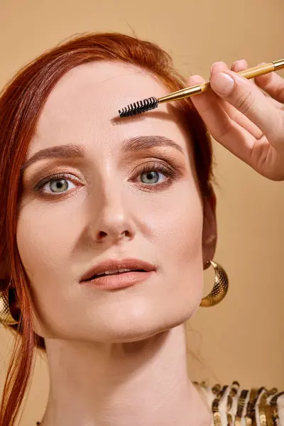 Porträt einer rothaarigen Frau mit grünen Augen mit Augenbrauenbürste auf beigem Hintergrund, Make-up — Stockfoto