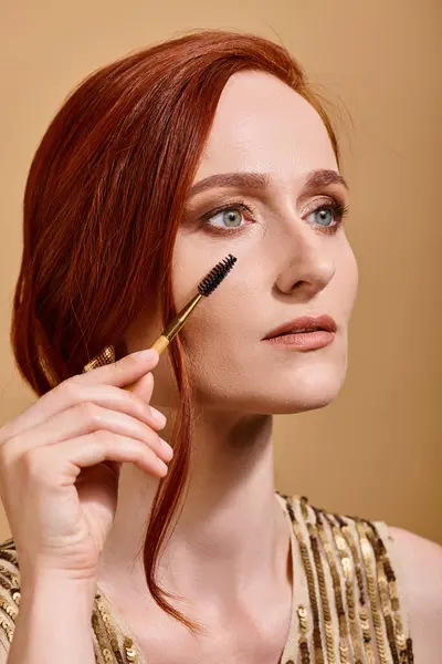Portrait of redhead woman with green eyes applying mascara on beige background, makeup beauty — Stock Photo