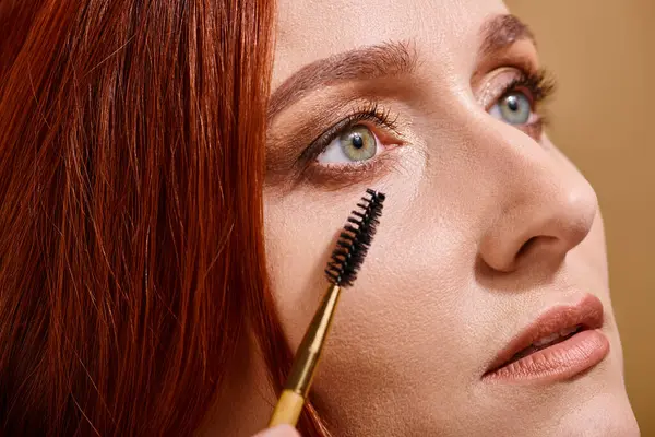 Nahaufnahme einer rothaarigen Frau mit grünen Augen, die Mascara auf beigem Hintergrund aufträgt, Make-up — Stockfoto