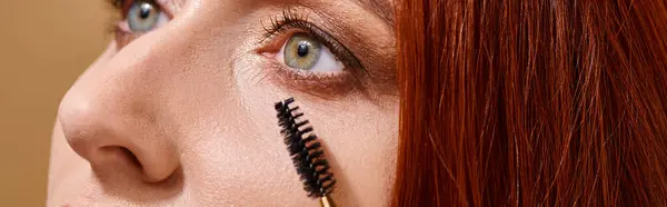 Abgeschnittenes Banner einer Frau mit grünen Augen, die Mascara auf beigem Hintergrund aufträgt, Make-up-Routine — Stockfoto