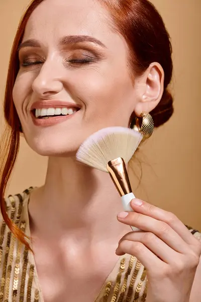 Happy redhead woman in gold sequin dress applying bronzer with makeup brush on beige background — Stock Photo
