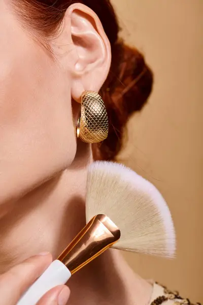 Cropped redhead woman in gold earring applying bronzer with makeup brush on beige background — Stock Photo