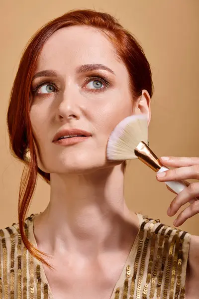Femme rousse rêveuse dans la trentaine appliquant bronzant avec pinceau de maquillage sur fond beige — Photo de stock