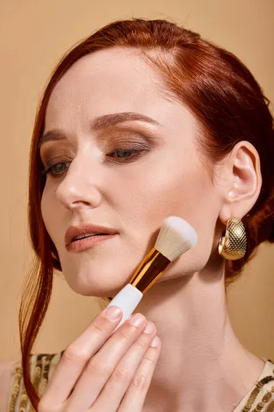 Redhead woman in gold earring applying face foundation with makeup brush on beige backdrop — Stock Photo