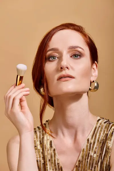 Confident redhead woman in gold earrings holding makeup brush on beige backdrop, beauty concept — Stock Photo