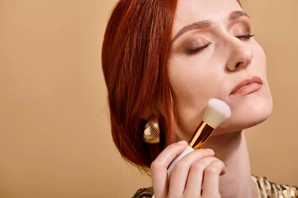 Redhead woman in gold earring applying face foundation with makeup brush on beige backdrop — Stock Photo